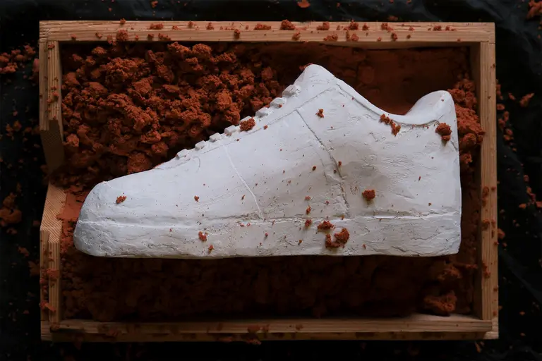 A white sneaker sculpture is placed on a bed of reddish-brown clay in a wooden box. The shoe appears textured, resembling a real sneaker, with some clay dust on its surface. The background is dark, emphasizing the shoe and the contrasting clay.