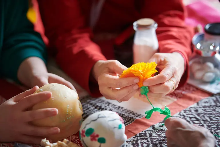 Zwei Hände halten einen Kunstblumenstrauß, während eine andere Hand eine bemalte Schüssel oder ein Gefäß berührt. Der Tisch ist mit bunten Stoffen und verschiedenen dekorativen Gegenständen bedeckt. Die Szene vermittelt eine Atmosphäre von kreativem Handwerken und gemeinschaftlichem Schaffen.