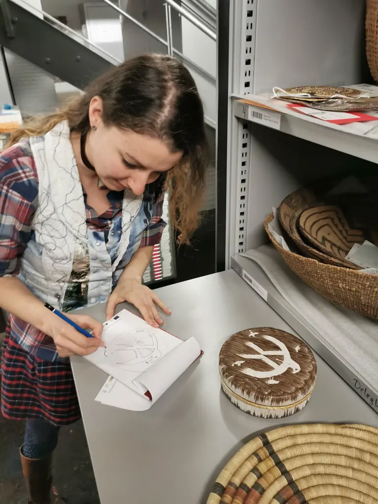 Eine Person steht an einem Tisch und zeichnet in ein Notizbuch. Neben ihr liegen verschiedene geflochtene Körbe und eine runde Schachtel mit einem dekorativen Muster. Im Hintergrund sind Regale sichtbar.
