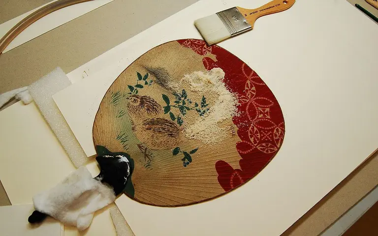 A traditional Japanese fan featuring a painted scene of quails and floral elements, surrounded by a red decorative border. Nearby are art supplies, including a paintbrush and tools for crafting. The background consists of light-colored materials used for mounting or presentation.