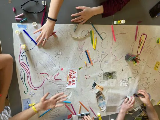 Eine Gruppe von Personen arbeitet an einem großen Papierblatt, das mit bunten Stiften, Kleber und anderen Bastelmaterialien bearbeitet wird. Einige Hände sind sichtbar, während verschiedene Zeichnungen und kreative Elemente entstehen. Der Fokus liegt auf dem kreativen Schaffensprozess und dem kollaborativen Arbeiten.