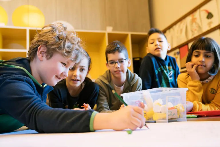 Eine Gruppe von fünf Kindern sitzt auf dem Boden und schaut aufmerksam zu, während eines von ihnen auf ein großes Blatt Papier zeichnet. Sie scheinen in einem kreativen Umfeld zu sein, umgeben von Materialien.
