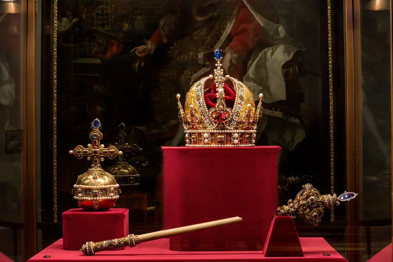 Eine goldene Krone mit roten und blauen Edelsteinen steht auf einem Samtpolster. Daneben sind ein Zepter und ein Reichsapfel, beide reich verziert. Die Objekte sind Teil einer Ausstellung historischer Krönungsinsignien.