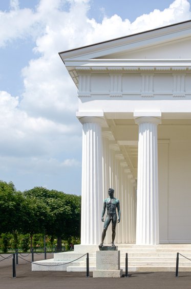Eine bronzene Statue eines stehenden Mannes befindet sich vor einem weißen, säulengestützten Gebäude. Der Hintergrund zeigt grüne Bäume und einen blauen Himmel mit einigen Wolken.