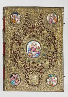 An ornate book cover featuring intricate silver filigree work, embellished with colorful enamel portraits of religious figures. The central oval depicts a seated mother and child, surrounded by smaller ovals depicting various scenes. The rich red background complements the ornate detailing.