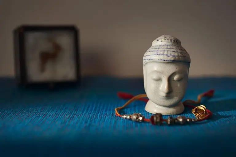 Eine kleine Buddha-Statue aus weißem Material steht im Vordergrund auf einem blauen Tuch. Im Hintergrund ist eine dekorative Kiste mit einem tierischen Motiv zu sehen. Vor der Statue liegt ein Armband mit verschiedenen Perlen und einem goldenen Element.
