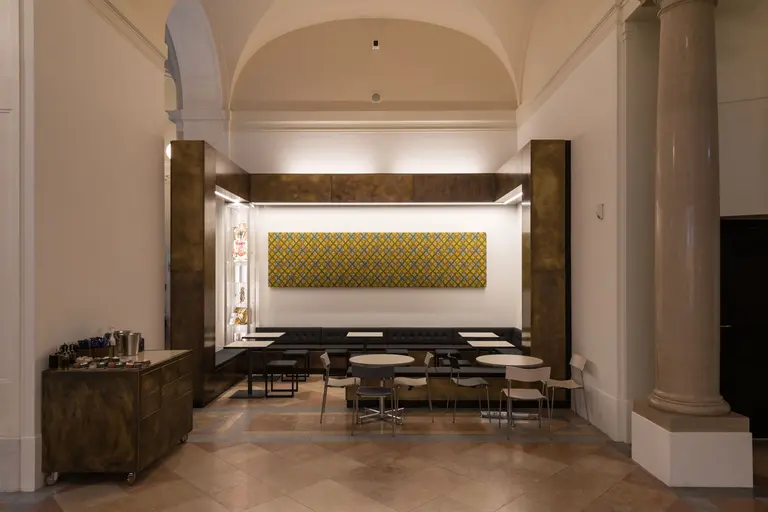 Der Raum zeigt einen modernen Sitzbereich mit eleganten Tischen und Stühlen. An der Wand hängt ein großes, geometrisches Kunstwerk in warmen Farben. Die Atmosphäre ist stilvoll und einladend, mit einem Fokus auf minimalistische Gestaltung.