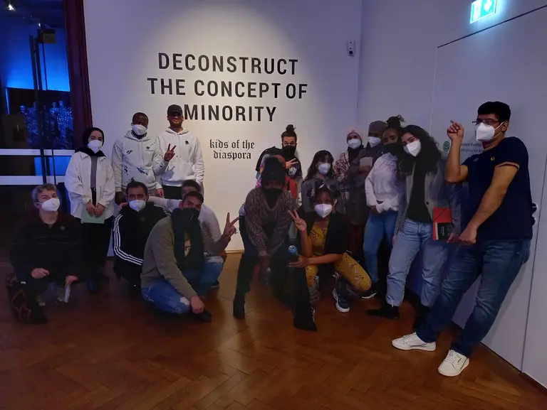Eine Gruppe von mehreren jungen Menschen posiert vor einer Wand mit der Aufschrift „DECONSTRUCT THE CONCEPT OF MINORITY“. Sie tragen Masken und zeigen verschiedene Gesten. Der Raum hat eine künstlerische Atmosphäre.
