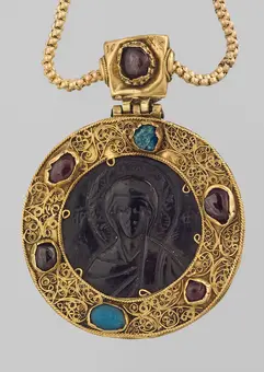 A round gold pendant featuring intricate filigree work, adorned with various gemstones including red and blue. At the center, there is a dark relief image of a figure, possibly religious. The pendant hangs from a gold chain.