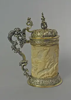 Decorative stein featuring an ornate metal handle and lid. The body is carved ivory, depicting intricate scenes of figures and animals. The lid is also elaborately designed, complementing the detailed craftsmanship of the stein.