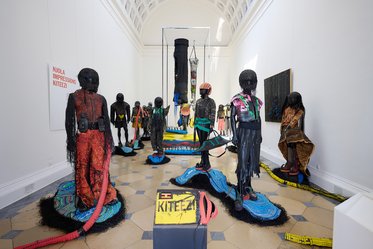 Eine Installation mit mehreren schwarzen Figuren aus verschiedenen Materialien in bunten Outfits. Sie stehen auf blauen und schwarzen Bodenplatten. Im Hintergrund sind weitere Kunstobjekte und eine große, hängende Struktur zu sehen. An der Wand hängt ein rotes Schild mit der Aufschrift "KITEZI".