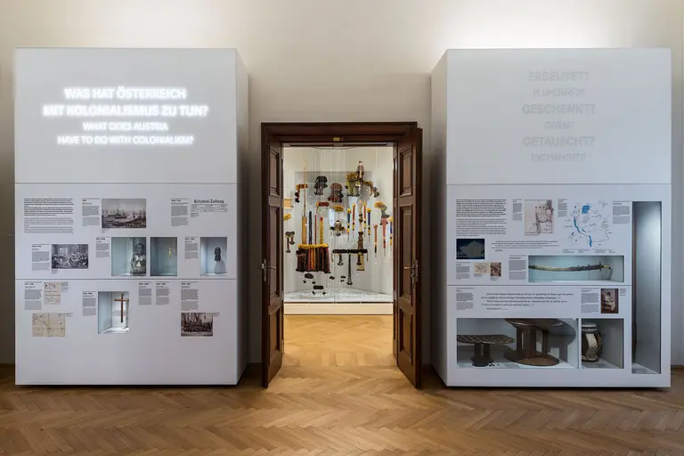 Ein Raum mit zwei beleuchteten Wänden, die Informationen über Österreichs Rolle im Kolonialismus bieten. An der gegenüberliegenden Wand ist ein Durchgang sichtbar, der in einen lebendig ausgestatteten Ausstellungsraum führt, der verschiedene Objekte zeigt.