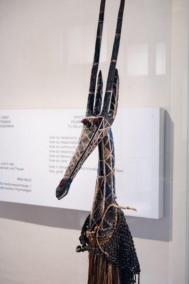 A tall, intricately designed sculpture resembling a gazelle, featuring elongated horns and decorative patterns. The neck is adorned with beads and other embellishments, standing against a blurred background that includes a display wall with text.