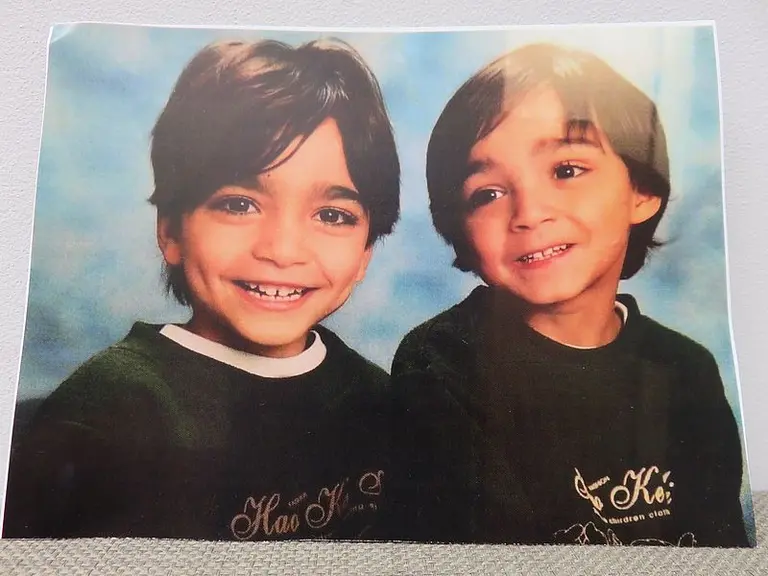 Zwei kleine Jungen lächeln in die Kamera. Sie haben dunkles Haar und tragen grüne Pullover mit Schriftzügen. Der Hintergrund ist blau und weich verschwommen, was den Fokus auf die Jungs lenkt.