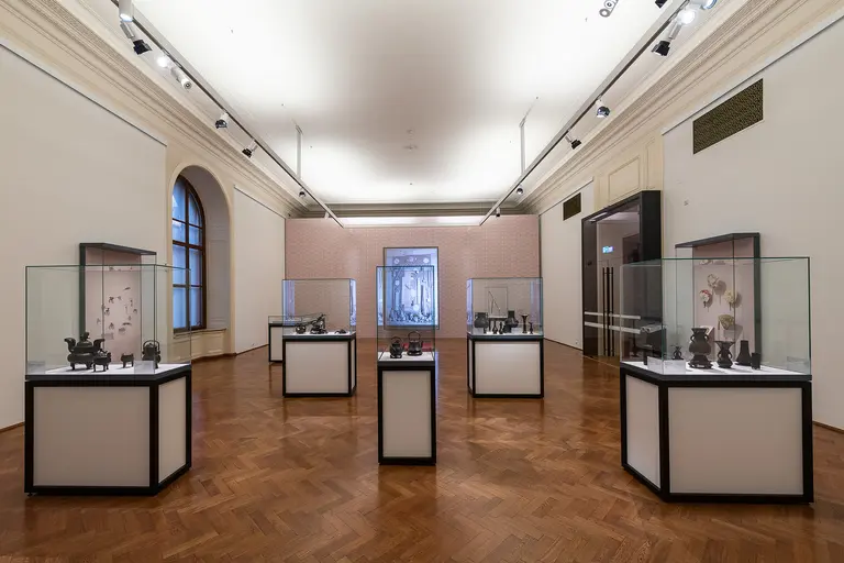 A spacious exhibition room features several display cases with artifacts inside. The cases are positioned on wooden flooring, and soft lighting highlights the items. A large backdrop artwork is visible in the center, adding context to the displayed pieces.
