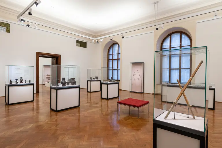 A spacious museum gallery featuring several glass display cases containing various artifacts. In the center, a red cushioned bench adds seating. Natural light filters through large windows, illuminating the wooden flooring and creating an inviting atmosphere for visitors.
