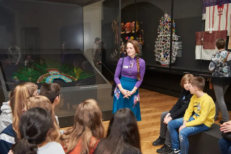 Eine Person mit langen, braunen Haaren steht vor einer Gruppe von Kindern und erklärt etwas. Im Hintergrund ist eine Vitrine mit bunten Kunstwerken und traditioneller Handwerkskunst zu sehen. Die Kinder hören aufmerksam zu und schauen in Richtung der Vortragenden.