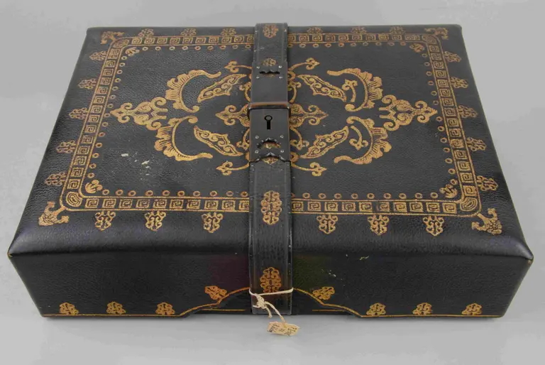A closed, rectangular box with a black leather exterior embellished with intricate gold designs. The box features decorative patterns and is secured with a strap and lock in the center.