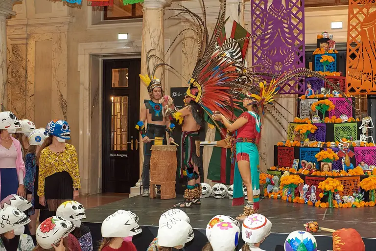 Auf einer Bühne führen Tänzer in bunten Kostümen mit Federn und traditionellen Motiven eine Feier auf. Im Vordergrund stehen Zuhörer in weißen Masken. Im Hintergrund ist ein farbenfrohes Altar-Arrangement mit Blumen und Figuren zu sehen, das festliche Stimmung vermittelt.