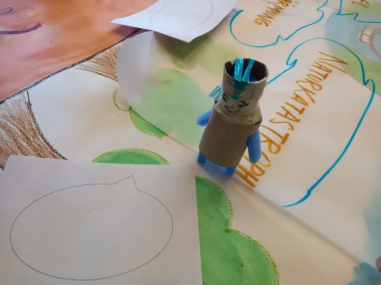 Eine bunte Bastelfigur aus Pappmaché steht auf einem Tisch, umgeben von großen, bemalten Papierbögen. Neben ihr liegen leere Blätter mit ovalen Umrissen, die möglicherweise für Zeichnungen oder Notizen genutzt werden sollen.