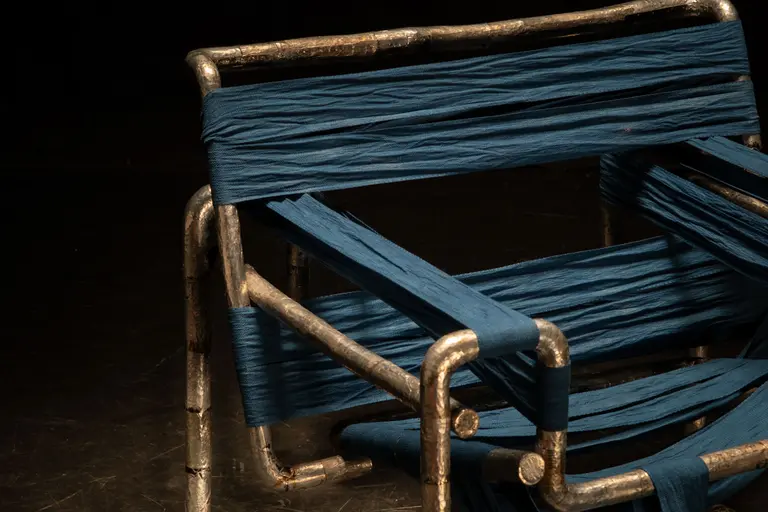 Ein Stuhl mit einem strukturierten, silbernen Metallrahmen, der mit blauen, texturierten Stoffstreifen umwickelt ist. Die Streifen verlaufen über die Armlehnen und die Sitzfläche, was dem Stuhl ein kreatives und modernes Aussehen verleiht.