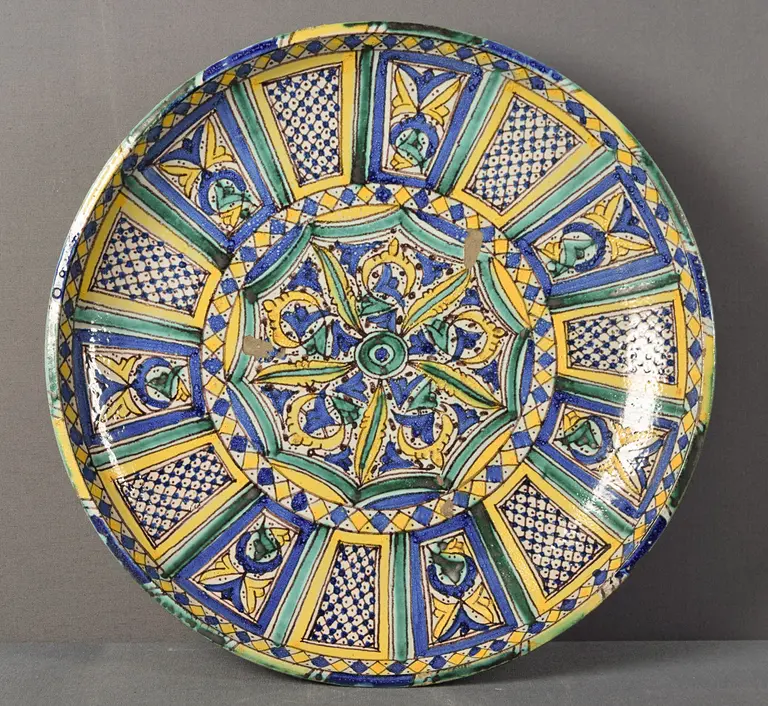 Ein großer, runder Teller mit detaillierten, bunten Mustern in Blau, Gelb und Grün. Im Zentrum befindet sich ein florales Design, umgeben von geometrischen Formen. Der Teller hat einen rustikalen, handgefertigten Look.
