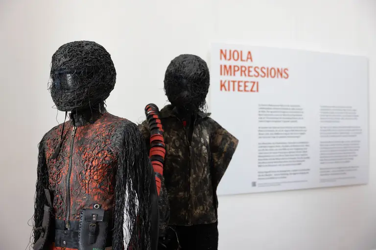 Zwei skulpturale Figuren stehen in einer Galerie. Die Figuren sind aus schwarzem Draht und Kleidungsstücken gefertigt, mit unterschiedlichen Texturen und Farben. Im Hintergrund befindet sich eine informative Wandtafel mit dem Titel „NJOLA IMPRESSIONS KITEEZI“.