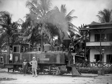 Eine historische Dampflok steht auf einem Gleis, umgeben von Palmen. Zwei Männer stehen davor, einer in uniformierter Kleidung und der andere in traditioneller Kleidung. Im Hintergrund sind Gebäude und weitere Personen zu sehen. Die Szenerie vermittelt einen kolonialen Einfluss.