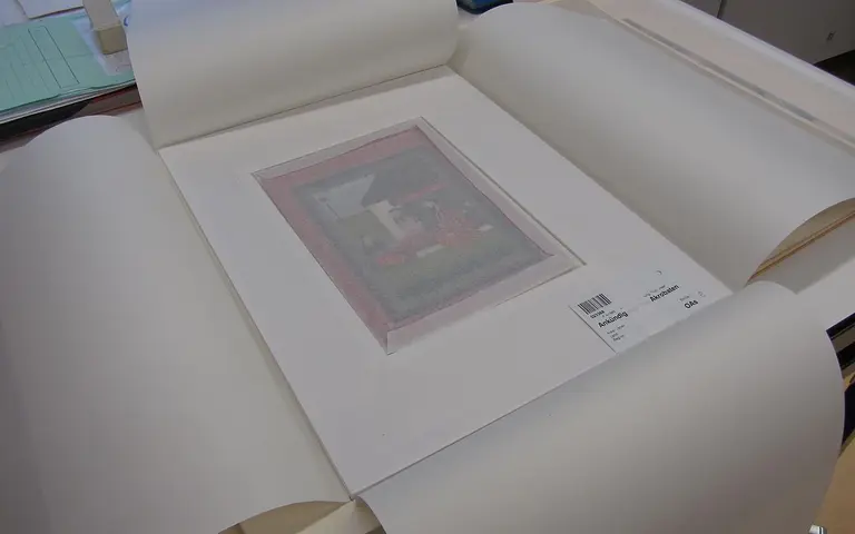 A framed artwork featuring a colorful illustration of a building and a vehicle is placed on a table. The artwork is surrounded by protective matting and wrapped in archival paper for preservation, with an identification label visible on the mat.
