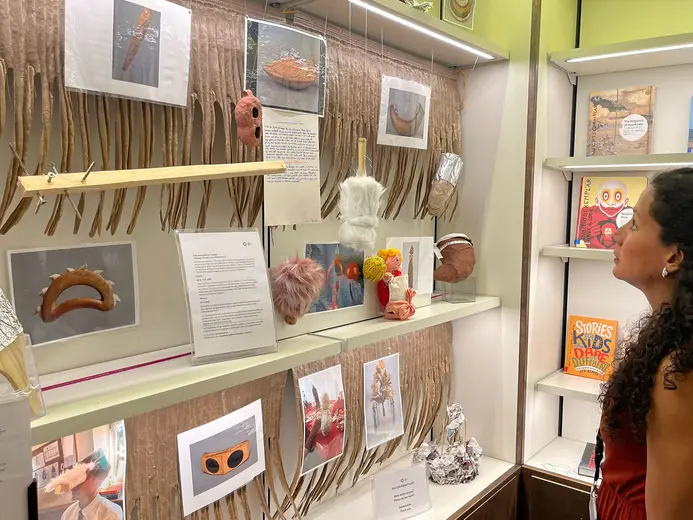 A woman examines a display featuring various artistic objects and masks, alongside descriptive text on a wall. The exhibit is decorated with textured materials, and there are photographs showcasing the items. Shelves in the background hold books and additional decorative items.