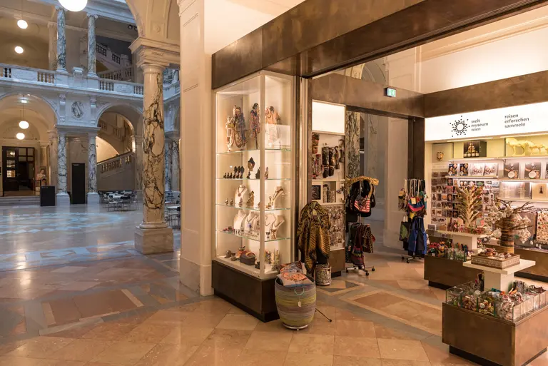 Ein Blick auf den Eingang eines Geschenkeladens in einem Museum. Rechts sind Regale mit Kunsthandwerk und Souvenirs zu sehen. Im Hintergrund erkennt man die architektonischen Details der Museumshalle mit bunten Marmoroberflächen und großen Lichtquellen.