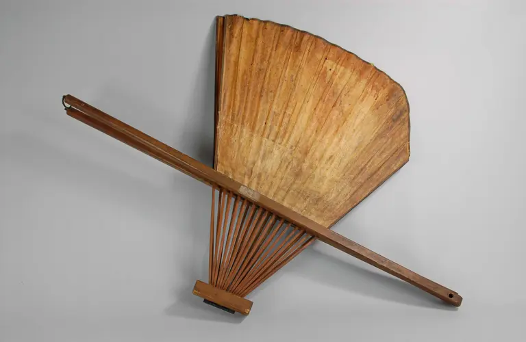 A large, decorative hand fan made of wood, featuring a wide, curved shape with slats radiating from a central handle. The fan is primarily brown, showcasing natural wood grain patterns. It is placed against a neutral gray background.
