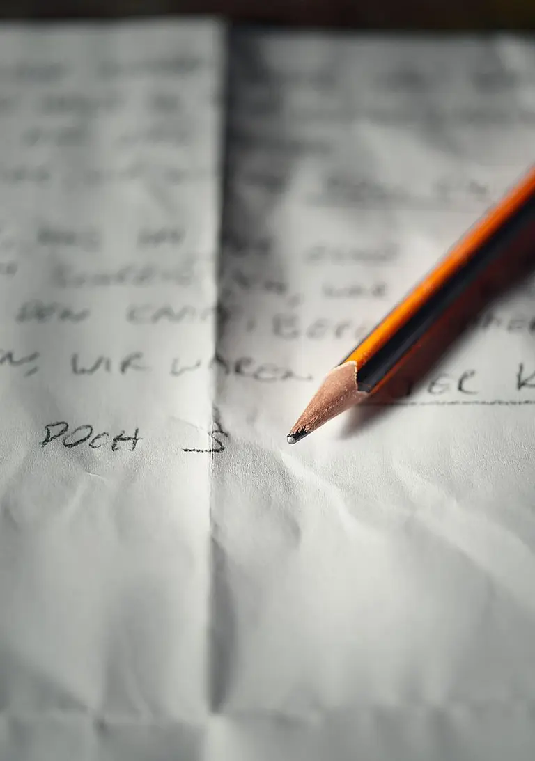 Ein zerknülltes Blatt Papier mit handschriftlichem Text liegt auf einem Tisch. Darauf lehnt ein orangefarbener Bleistift, dessen Spitze zum Schreiben bereit ist. Die unscharfen Worte auf dem Papier scheinen eine persönliche Botschaft zu vermitteln.