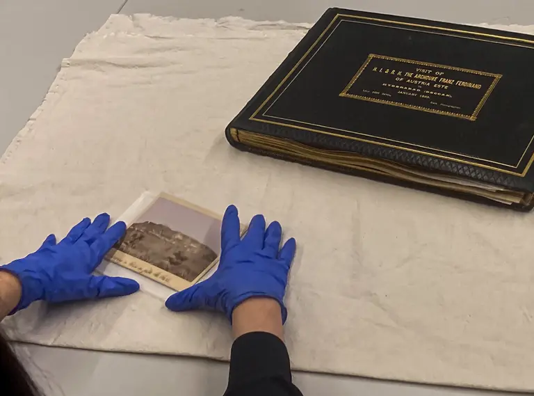 Eine Person mit blauen Handschuhen liegt vor einem Album auf einem Tisch. Sie hält ein vergrößertes Foto mit einer historischen Ansicht. Das Album hat einen schwarzen Einband mit goldener Schrift und liegt neben einem weißen Tuch.