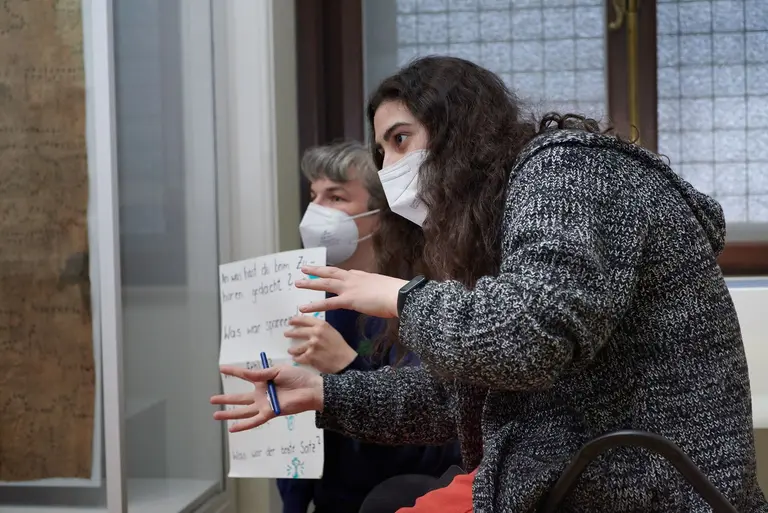 Zwei Personen tragen Masken und sitzen in einem Raum. Eine Person gestikuliert und hält einen Stift, während sie spricht. Die andere Person schaut aufmerksam zu und hält ein Plakat mit Text. Das Bild vermittelt eine Diskussion oder Präsentation.