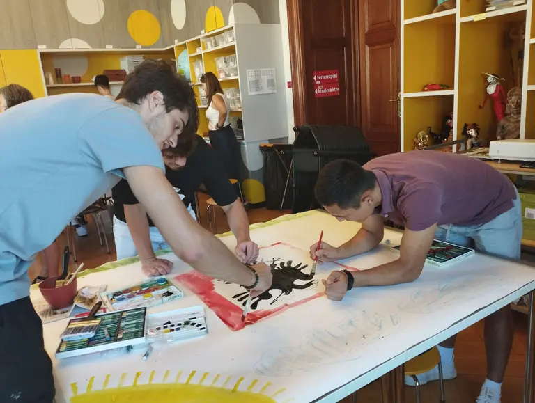 Drei junge Männer arbeiten an einem großen Kunstwerk auf einem Tisch. Sie malen mit Farben auf eine Leinwand und konzentrieren sich auf ihre Gestaltung. Im Hintergrund sind mehrere Personen und ein kreativer Raum mit bunten Wänden und Regalen sichtbar.