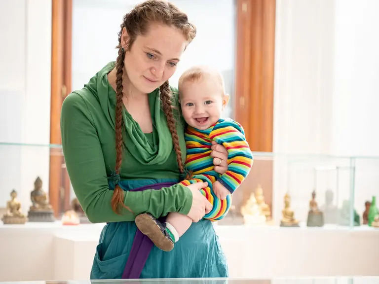 A woman with long, braided hair holds a smiling baby in a colorful striped outfit. They are in a bright indoor space, with display cases containing small statues in the background. The woman wears a green top and a blue skirt, looking affectionately at the baby.