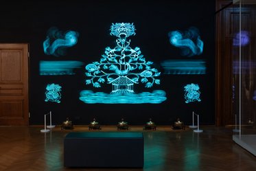 A dark room features a large, illuminated blue artwork on the wall, depicting a stylized tree surrounded by floral motifs. Below, small projection devices emit soft light, creating an ethereal atmosphere. A dark bench is positioned in the foreground for viewers to sit and observe.