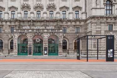 The image shows the entrance of the Welt Museum Wien in Vienna. The building features ornate architecture with three large arched green doors. Above the entrance, the museum's name is displayed in white letters. A rectangular black structure stands beside the entrance, and the ground is covered with a reddish-orange pathway.