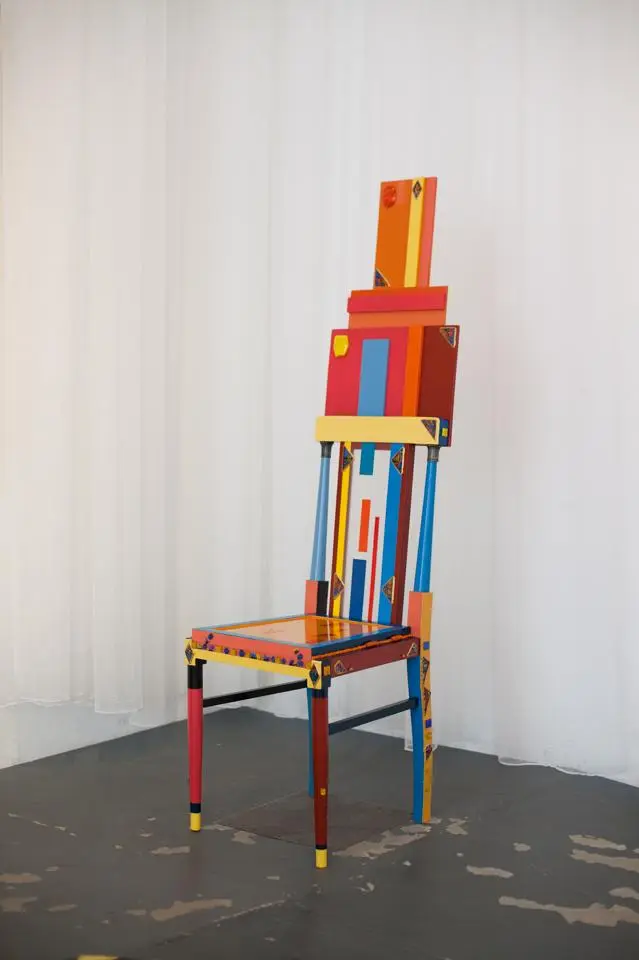 Ein farbenfroher Stuhl mit geometrischen Mustern steht auf einem harten Boden. Der Stuhl hat eine hohe Rückenlehne, die kreativ gestaltet ist und aus verschiedenen farbigen Elementen besteht, die in vertikalen und horizontalen Linien angeordnet sind.