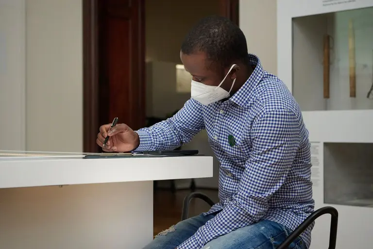 Ein Mann mit einer Gesichtsmaske sitzt an einem Tisch und schreibt mit einem Stift. Er trägt ein kariertes Hemd und Jeans, und ist in einem Innenraum zu sehen, möglicherweise in einem Museum oder einer Galerie.