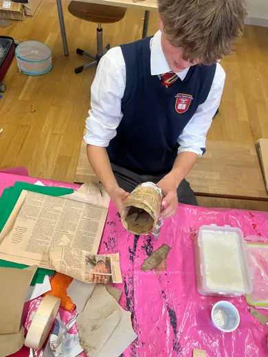 A student in a school uniform is working on an arts and crafts project, holding a paper tube and surrounded by various craft materials, including newspaper and cardboard pieces. The workspace features a bright pink table covering and other crafting supplies.