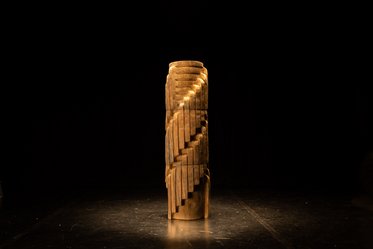 Eine skulpturale Holzsäule mit spiralartigen Vertiefungen und einer eleganten Form steht im Mittelpunkt auf einer dunklen Bühne. Die Beleuchtung hebt die Struktur und Textur des Holzes hervor.