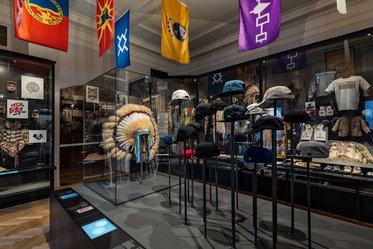 A museum display featuring a variety of hats on stands, including traditional Native American headdresses. Behind the hats, colorful flags hang, and artifacts are visible in glass cases, showcasing cultural heritage and history. The setting provides an educational experience about indigenous cultures.