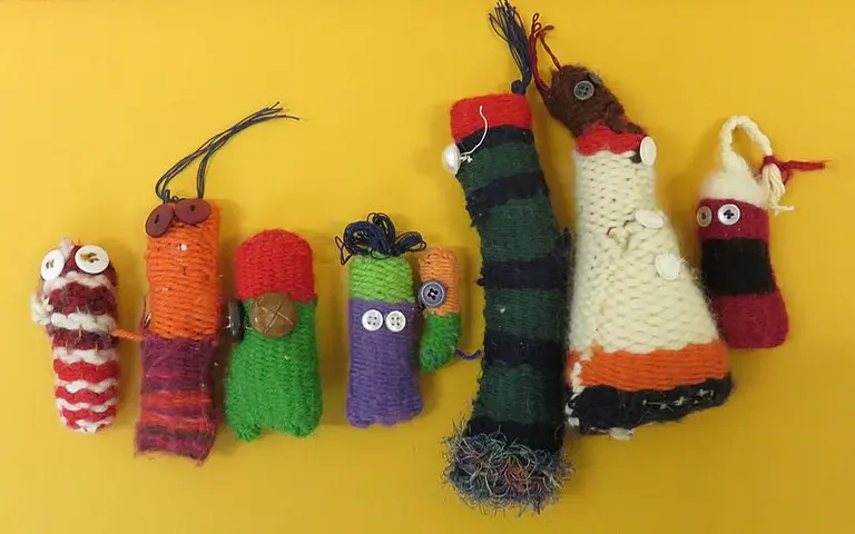 Bunte, handgefertigte Figuren aus Garn, die in verschiedenen Größen und Farben geformt sind. Einige haben Knöpfe als Augen, während andere unterschiedliche Muster und Texturen aufweisen. Die Figuren stehen in einer Reihe vor einem gelben Hintergrund.