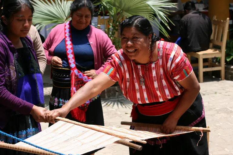 Drei Frauen in traditioneller Kleidung arbeiten zusammen an einem Webrahmen. Eine Frau lächelt fröhlich, während sie den Webstuhl bedient. Die Umgebung ist hell und freundlich, mit Pflanzen im Hintergrund. Die Szene zeigt eine lebendige, handwerkliche Aktivität und Gemeinschaft.