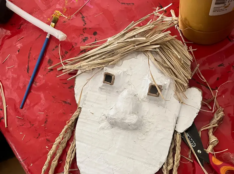 A child's craft project featuring a face made from cardboard. The face has straw for hair, two cut-out eyes, and features like a nose and mouth. Scissors and a glue bottle are visible in the surrounding area, on a red table surface.