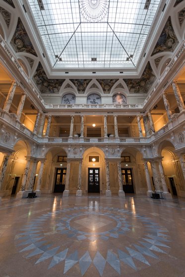 A grand interior view of a historical building featuring ornate marble columns and intricate architectural details. Natural light floods in through a large skylight, illuminating the spacious area adorned with a circular floor pattern and several doorways leading outside.