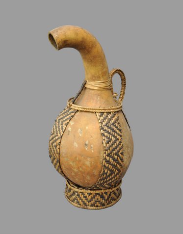 A decorative, woven vessel with a long, curved spout and a rounded body. The body features intricate patterns created from natural materials, showcasing traditional craftsmanship. The vessel is designed to hold liquid and is displayed against a plain gray background.
