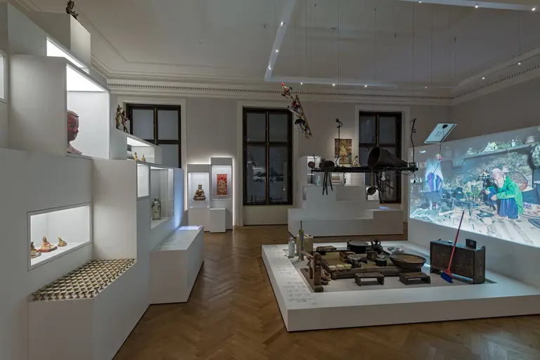 Das Bild zeigt eine Ausstellungsfläche mit verschiedenen Kunstwerken und Objekten. An den Wänden hängen Bilder, während auf einem Tisch historische Gegenstände präsentiert sind. Eine Projektion auf der Wand zeigt eine Szene mit Menschen. Die Räumlichkeit vermittelt eine moderne, künstlerische Atmosphäre.